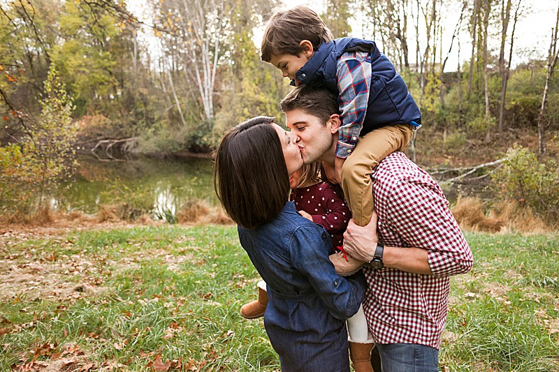 Mom and dad kissing with their toddler son on dad's shoulders.