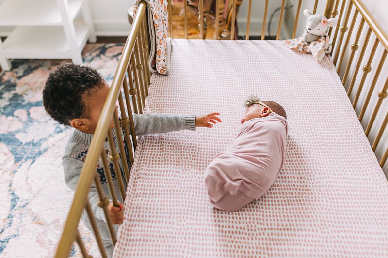Toddler reaching into a crib to touch his newborn sister.